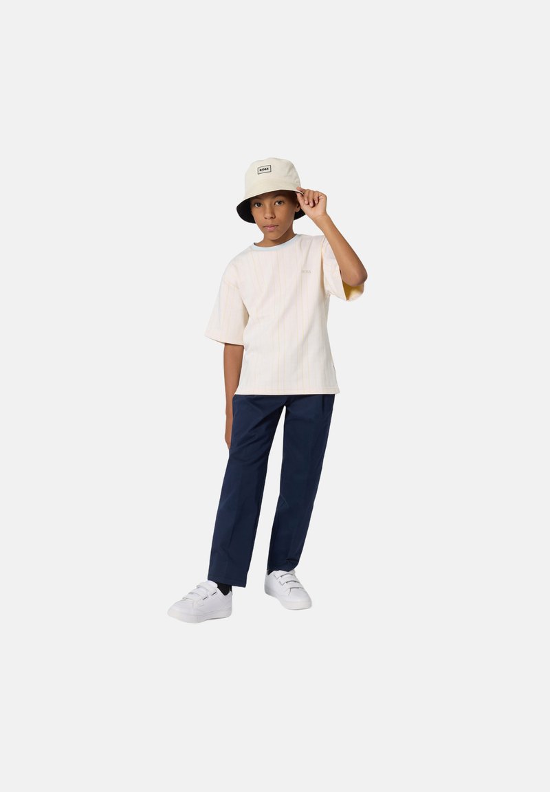 Jeune portant une chemise à manches courtes à rayures légères, un pantalon marine et des baskets blanches ; porte également un chapeau seau beige avec une bordure noire.