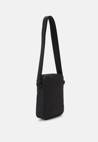 Czarna prostokątna torba na ramię z długim, regulowanym paskiem, prezentowana na tle jasnej, białej ściany.