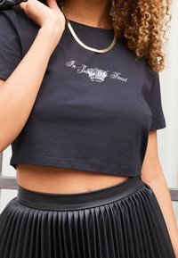 T-shirt noir court en coton, avec le texte blanc "In Just Trust" accompagné d'un graphique de couronne. Associé à une jupe en cuir plissée noire.