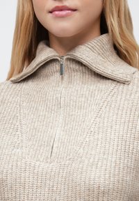 Pulover bej tricotat cu guler înalt, prevăzut cu fermuar pe sfert. Materialul are un model texturat, cu dungi și un luciu subtil.