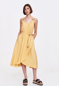 Żółta sukienka bez rękawów z dekoltem w kształcie litery V, regulowanymi ramiączkami, wcinającą talią i asymetrycznym dołem. Połączona z czarnymi sandałami.