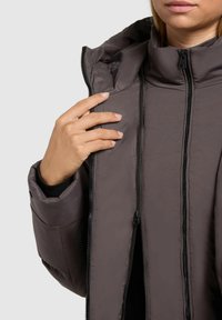 Isik kannab tumedat halli polsterdatud jope, millel on kaks nähtavat lukku, hoides kraed ja osaliselt avades jope.