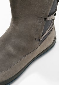 Grå mockaboot med rundad tå, kontrasterande svart panel och dekorativa snörningsdetaljer längs sidan. Flat sula i gummi.