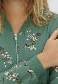 Zielona kurtka typu bomber z kwiatową haftem w białym i różowym kolorze. Posiada zamek błyskawiczny z przodu oraz ściągacze przy mankietach. Gładka faktura i dopasowany krój.