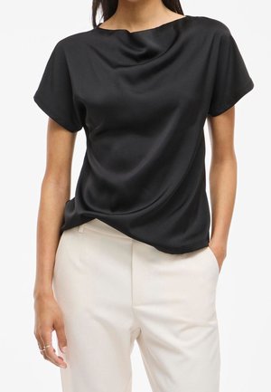 Kobieta w krótkim rękawie czarnej satynowej bluzce włożonej do białych spodni z wysokim stanem, jedna ręka w kieszeni, jednolite tło.