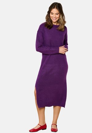 Robe tricotée à manches longues, couleur violet, avec des fentes sur les côtés, longueur mi-mollet. Le modèle l'a associée à des ballerines rouges, mettant en avant une coupe décontractée et une texture douce.