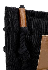 Sort vævet taske med en tekstureret overflade, der har en brun læderdetalje og et snoet sort reb ved lukningen.