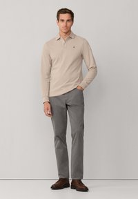 Polo beige à manches longues avec col, chinos gris, et chaussures en cuir marron. Présente un petit logo sur la poitrine. Design simple.