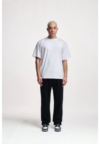 Vit, oversized bomull t-shirt i kombination med svarta byxor. Personen står rak med svarta och vita sneakers, och har kort ljus hår.