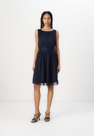 Femeie în rochie bleumarin fără mâneci, până la genunchi, cu talie mulată și sandale negre cu toc înalt și barete, pe un fundal alb simplu.