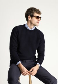 Marineblauwe trui met een gestructureerd breipatroon, gelaagd over een lichtblauw geruit shirt. Model draagt een bruine zonnebril en zit op een houten kruk.