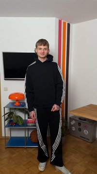 Young man with tattoos wearing black tracksuit with white stripes stands in modern room with shelving, orange lamp, basketball, and TV.