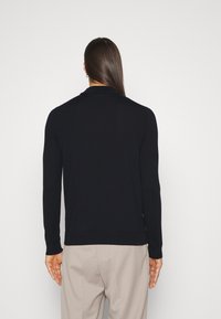Cardigan tricot à manches longues bleu marine avec col, doté d'une texture douce et de poignets côtelés, associé à un pantalon beige clair.