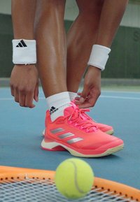 Lyse rosa sportssko med sølvdetaljer, flat såle og mesh-materiale, kombinert med hvite håndleddsbånd på solbrune armer, på en tennisbane.