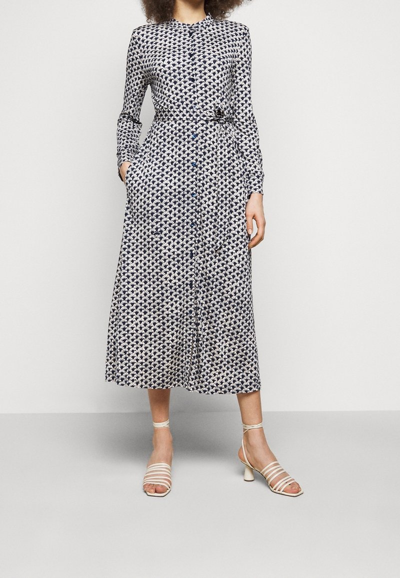 Femme portant une robe midi à manches longues, à motifs noir et blanc, avec des poches, associée à des sandales à talons beiges à brides.