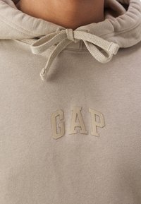 Beige mikina s texturovanou tkaninou, s kapucí, šňůrkou a vyvýšeným, světle bežovým logem "GAP" na přední straně.