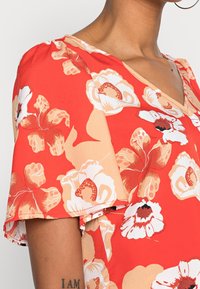 Blusa roja con flores que presenta un escote en V, mangas cortas con volantes, y una mezcla de patrones botánicos en crema y durazno sobre una tela texturizada.
