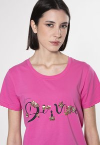Camiseta de algodón rosa con texto en negro en estilo de escritura, adornada con diversos acentos metálicos y joyas en la parte delantera.