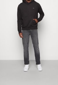 Felpa nera con cappuccio e tasca frontale, jeans grigi e sneakers bianche. La felpa presenta un piccolo logo sul petto.
