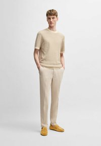 Beige struktureret strik t-shirt med korte ærmer, parret med lyse beige bukser og gule ruskind loafers, model står med hænderne i lommerne.
