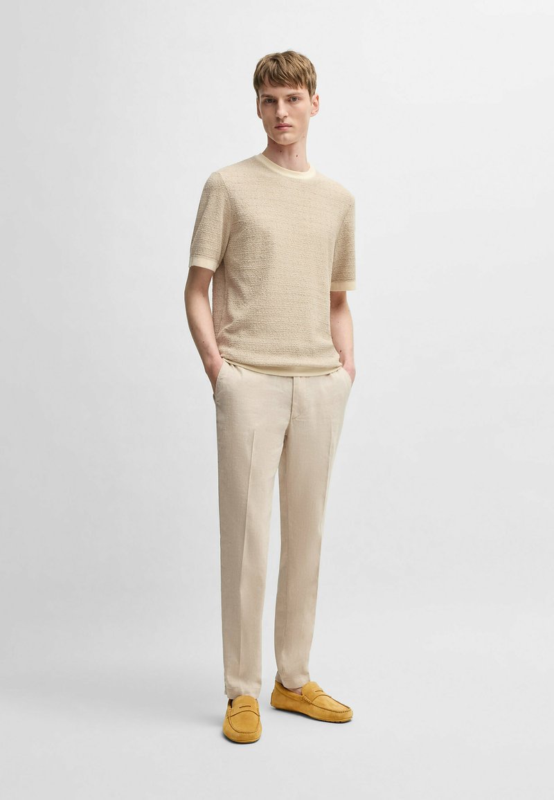 Beige struktureret strik t-shirt med korte ærmer, parret med lyse beige bukser og gule ruskind loafers, model står med hænderne i lommerne.