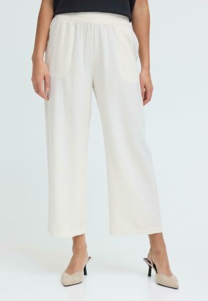 Una persona indossa pantaloni bianchi larghi a vita alta con elastico in vita e décolleté slingback beige con punta appuntita, in piedi contro uno sfondo semplice.