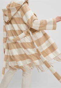 Osoba v béžovém a bílém kostkovaném kabátu s kapucí a třásněmi, s páskem v pase, zobrazená ze zad a ze strany.