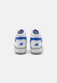 Paire de baskets montantes New Balance blanches et bleues vues de dos sur un fond clair uni.