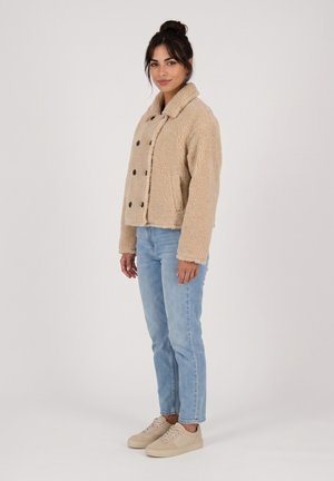 Mujer con una chaqueta beige peluda, vaqueros azules claros y zapatillas beige, de pie contra un fondo claro y liso.
