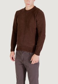 Maglione a maglia marrone con collo rotondo e maniche lunghe, con polsini e orlo a costine. Indossato sopra pantaloni grigi, che mostrano una finitura texturizzata.