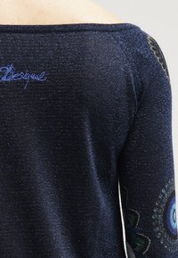 Dunkelblauer Pullover mit glitzernder Textur, schulterfreier Gestaltung und bunten gemusterten Akzenten an den Ärmeln. Blaues gesticktes Logo.