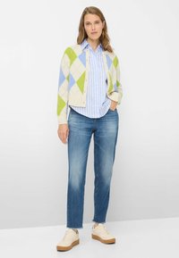 Gestrickte Strickjacke in Creme mit grünem und blauem Rautenmuster, über einem hellblau gestreiften Hemd getragen. Kombiniert mit hellblauen Jeans und beigen Sneakers.