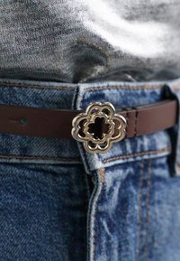 Pruun nahkade vöö, millel on lillekujulise hõbedase klambriga detail, kantud siniste denimste viisidega, millel on oranž õmblus.
