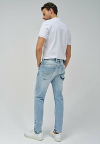 Jeans de mezclilla azul claro con un corte ajustado, con costuras amarillas y dos bolsillos traseros, combinados con una camisa polo blanca y zapatillas blancas.