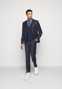 Mornarski trodijelni odijelo slim kroja, teksturirane tkanine, uz odgovarajuću kravatu. Bijele tenisice upotpunjuju izgled, naglašavajući ležernu eleganciju.