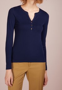 Marinblå långärmad tröja med V-hals och tre gyllene knappringar, tillverkad av mjukt material. Kombinerad med beiga skräddarsydda byxor.