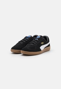 Schwarze Wildledersneaker mit weißen Seitenstreifen, Gummirutsche, runder Zehenform, flachen Schnürsenkeln und blauen Markenakzenten auf der Zunge.