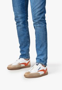 Sneaker bianchi e beige con accenti arancioni, dotate di una finitura in suede testurizzato, lacci piatti e suole in gomma, abbinate a jeans blu.