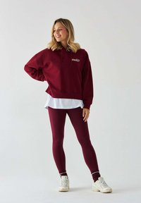 Bordowa hoodie z białym logo, nałożona na białą koszulę, zestawiona z dopasowanymi legginsami i białymi butami sportowymi.