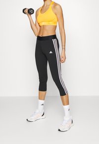 Donna in leggings nere Adidas e reggiseno sportivo giallo che solleva un manubrio da 3 kg, indossando calze e sneakers bianche Adidas su uno sfondo bianco.