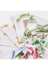En spridd uppställning av ansiktsmasker med olika botaniska illustrationer, inklusive ingredienser som aloe, honung och frukter på strukturerat papper.