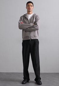 Lys grå zip-up hettegenser over en hvit skjorte, kombinert med svarte vide bukser og svarte sneakers. Jevn stoff med en avslappet passform.