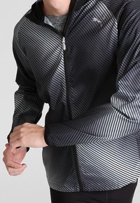 Giacca sportiva a strisce nera e grigia con chiusura a zip, dotata di cappuccio e realizzata in materiale liscio e leggero. Logo in alto a sinistra.