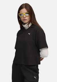 Sort kortærmet polo skjorte med et tekstureret mønster og et hvidt Puma logo. Laget med en hvid turtleneck nedenunder. Gule solbriller.