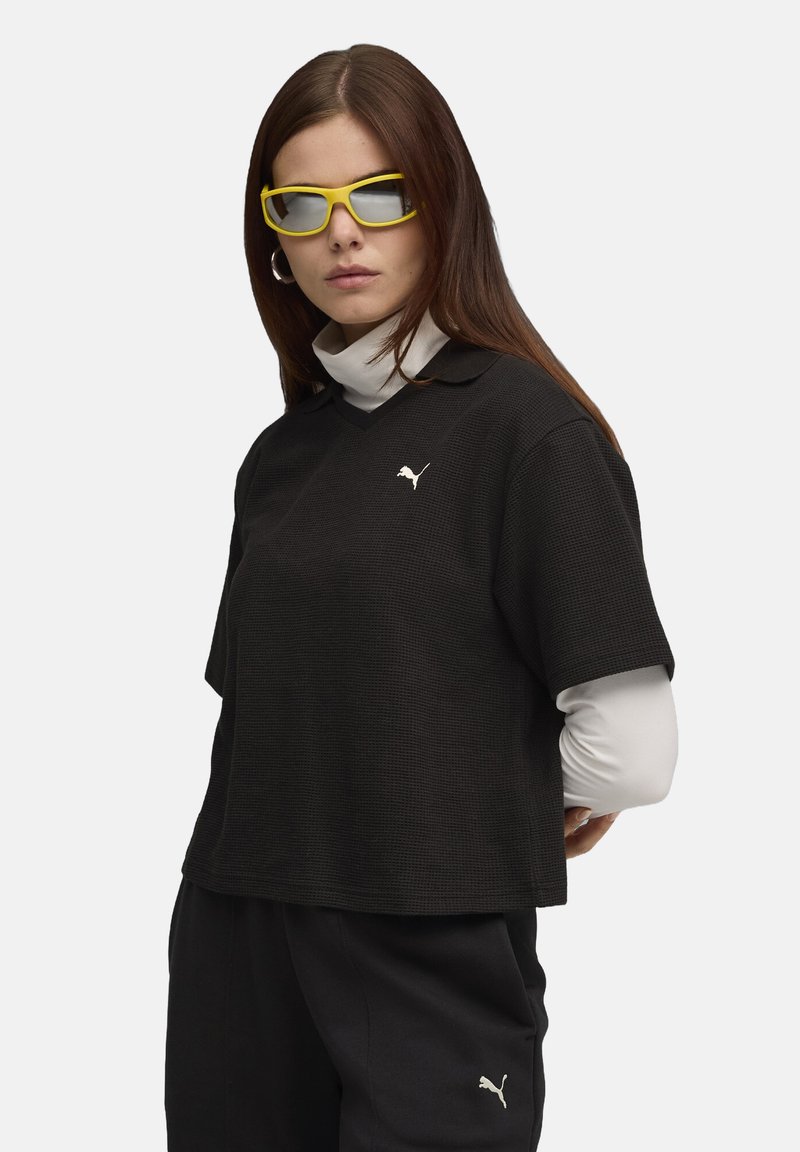 Sort kortærmet polo skjorte med et tekstureret mønster og et hvidt Puma logo. Laget med en hvid turtleneck nedenunder. Gule solbriller.