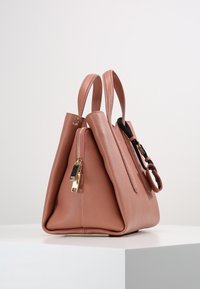 Vaaleanpunainen teksturoitu nahkalaukku, jossa on kaksoiskahvat, kultaiset vetoketjut ja musta yksityiskohta hihnassa, esillä valkoisella jalustalla.