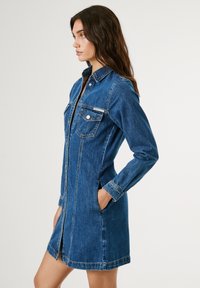 Robe en denim d'un bleu moyen avec un col, des manches longues, des boutons sur le devant et deux poches à la poitrine. Présente une silhouette en A flatteuse avec des poches latérales.