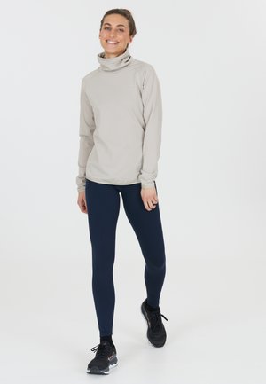 Helles beige Langarmoberteil mit hohem Kragen, kombiniert mit marineblauen Leggings und schwarzen Sportschuhen. Glatte Textur, figurnahe Passform.