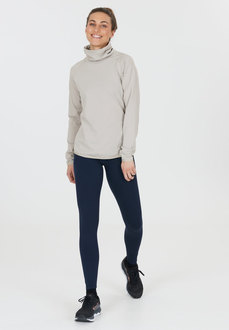 Lys beige langærmet top med høj krave, sammen med navy blå leggings og sorte træningssko. Glat tekstur, tætsiddende design.