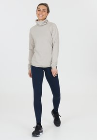Hellgraues Langarmoberteil mit stehendem Kragen, kombiniert mit marineblauen Leggings. Das Material hat eine glatte Textur; das Outfit ist für aktive Freizeitgestaltung konzipiert.